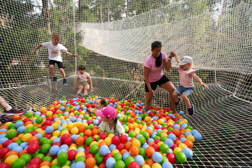 Lapset ja nainen seikkailevat puihin kiinnitetyssä verkossa, verkossa näkyy värikkäitä palloja